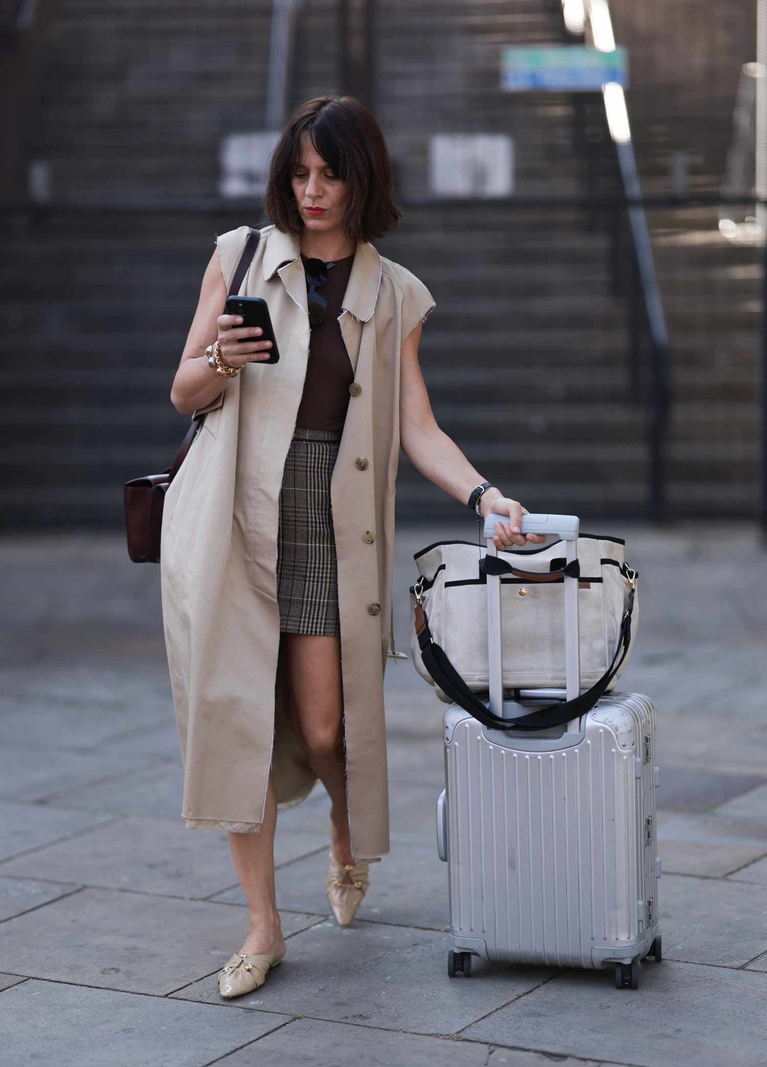 Woman in sleeveless trench coat rolls a suitcase down the street