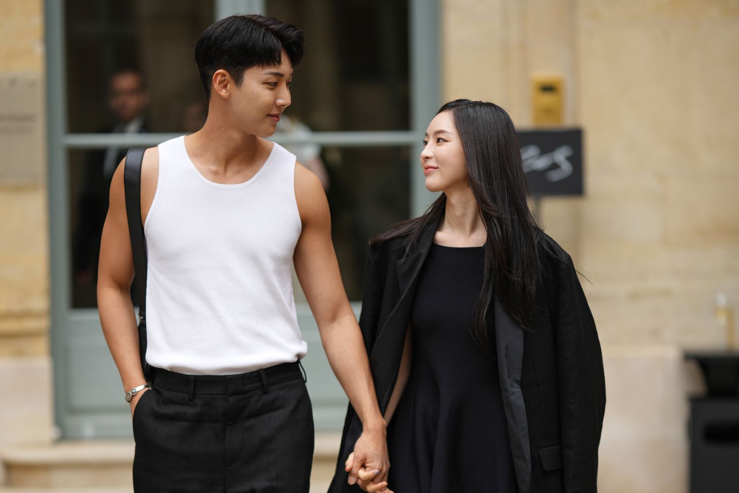 Jeongjin Park wears a white tank-top, a black shiny leather shoulder bag, black suit shorts, a silver bracelet, ; Sera Song wears silver earrings, a black short dress, a black blazer jacket, outside the COS show, on April 26, 2023 in Paris, France