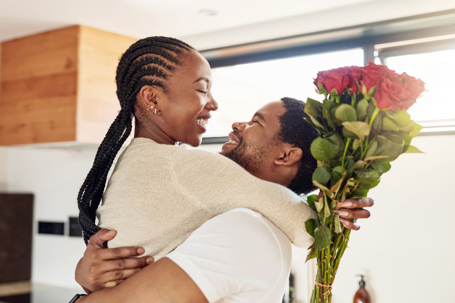 A couple smiles at each other and hugs. The woman holds a bouquet of red roses,