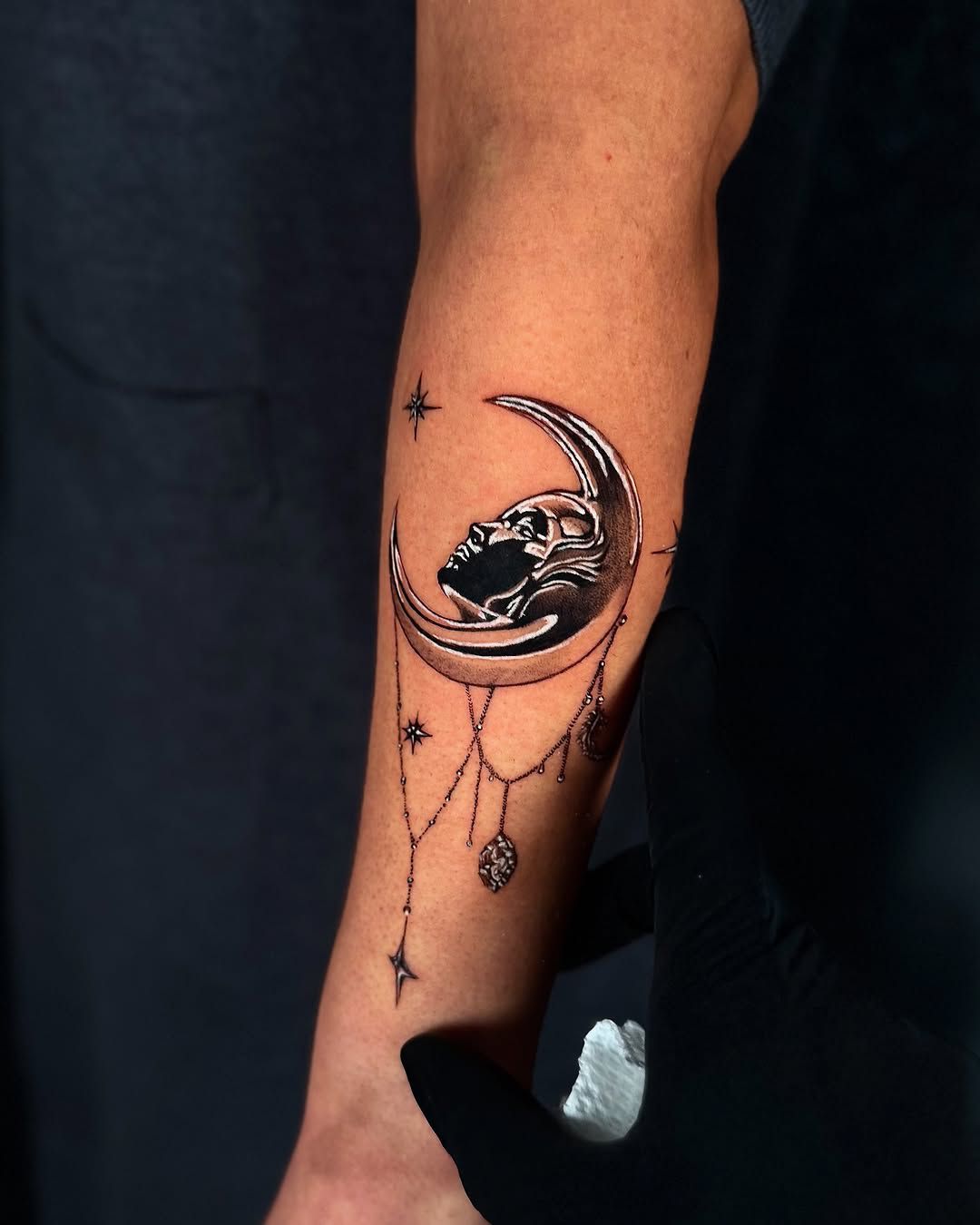 A metallic moon, with a face gently resting in it and jewelry chains hanging beneath it