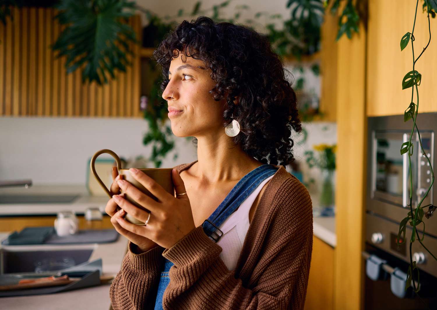 Person holding a mug, standing in a kitchen, gazing to the side amidst plants and wooden cabinetry.