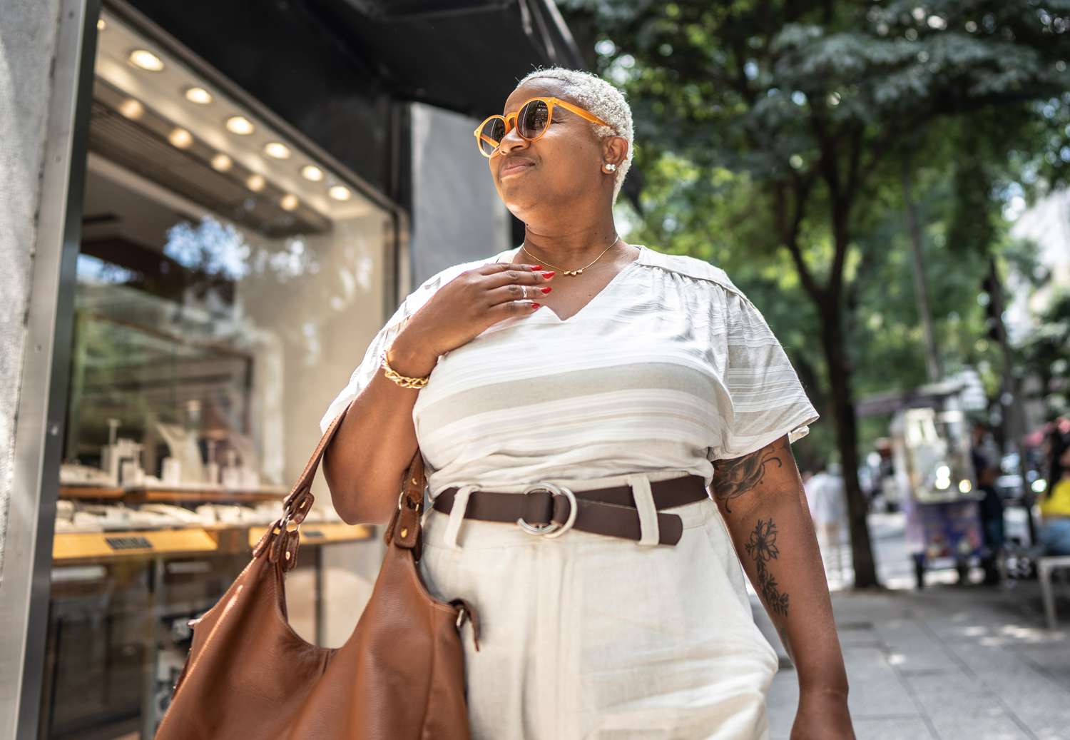 A person standing confidently outdoors, wearing sunglasses and holding a bag, with a jewelry store window in the background