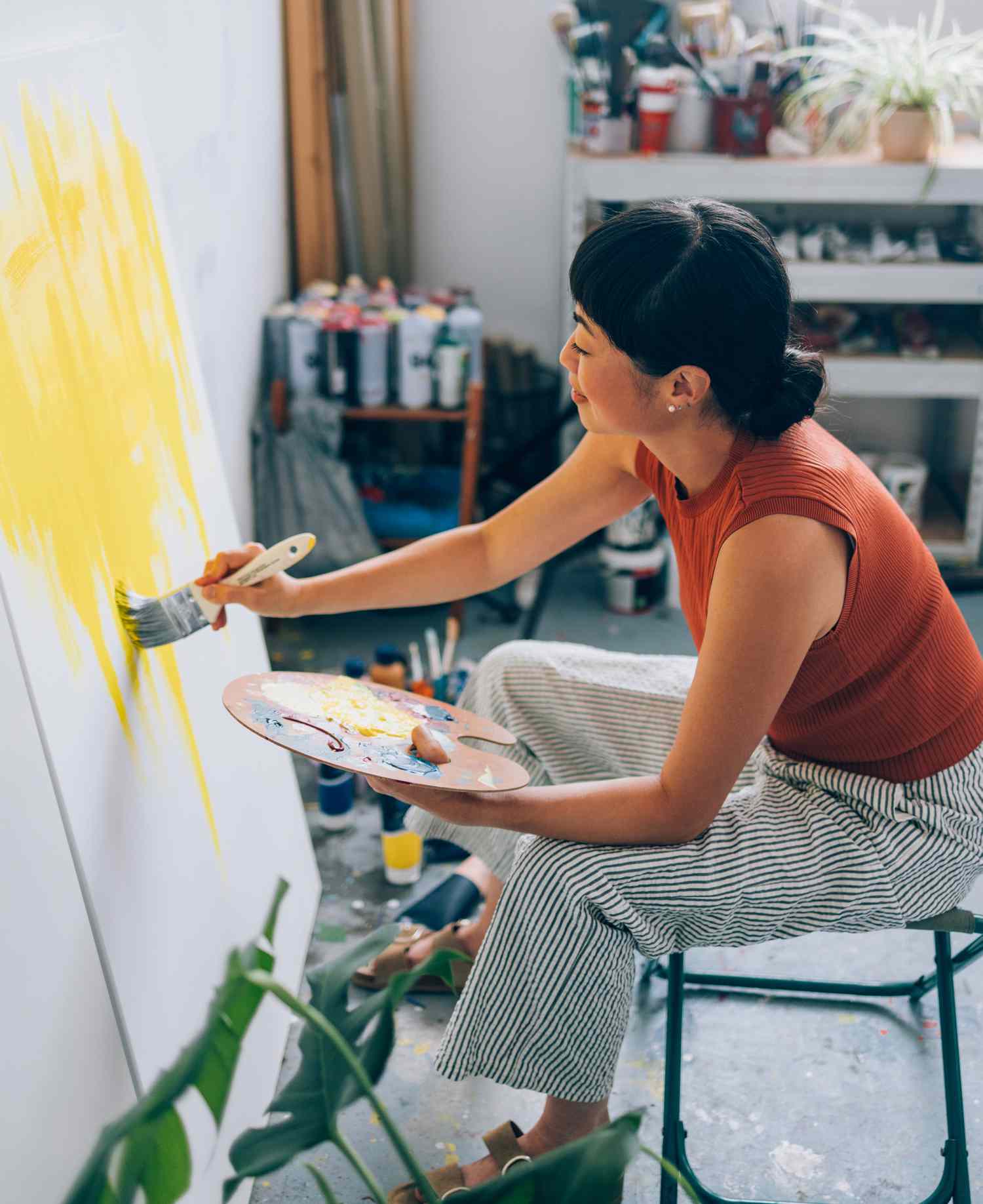 A person painting abstract yellow strokes on a canvas in an art studio, holding a brush and palette