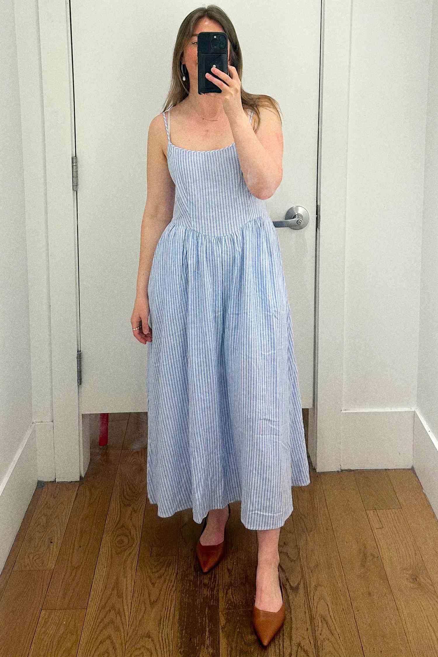 A person taking a mirror selfie wearing a long dress and heels in a fitting room, holding a phone to obscure their face