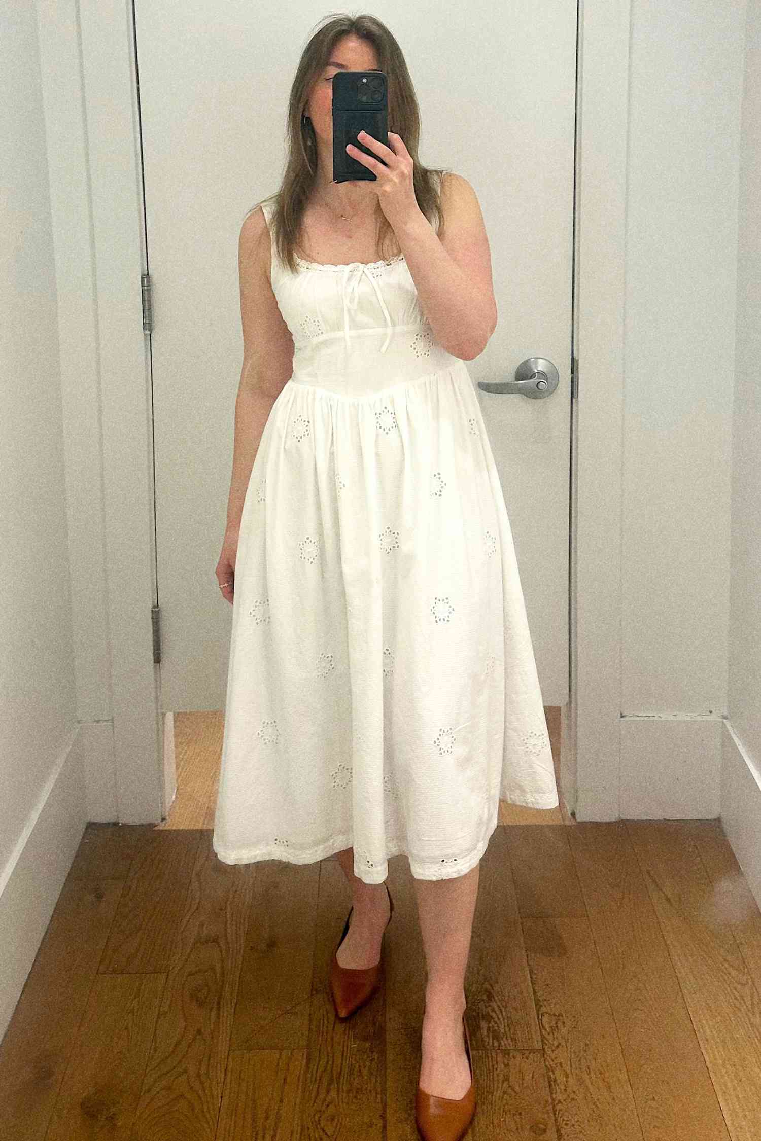 A person taking a selfie in a dressing room while wearing a knee-length white dress and flats