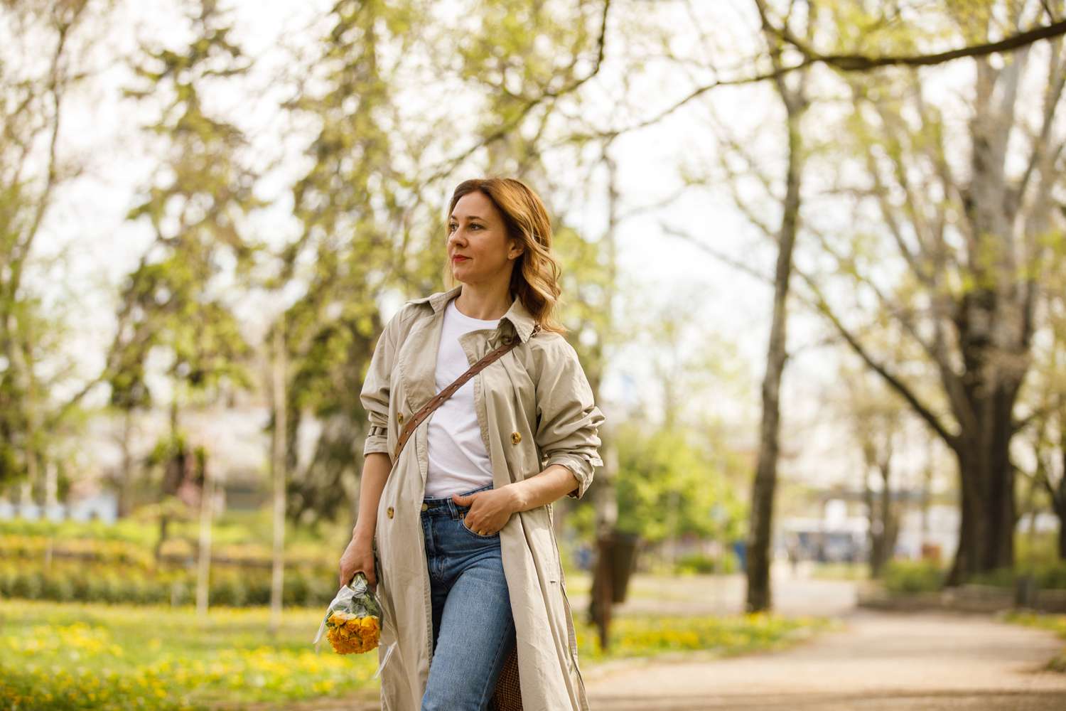 Adult blond woman looking away during a relaxing walk at the park carrying a bouquet of yellow roses.
