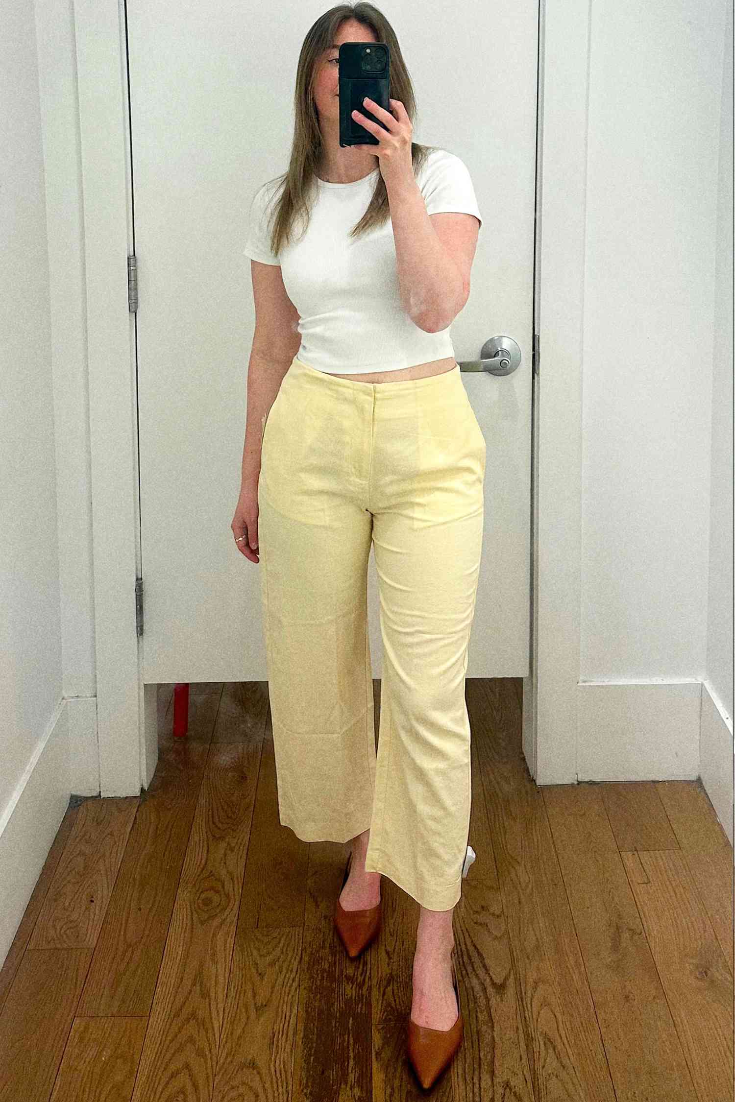 A person taking a mirror selfie wearing a white top and yellow pants, standing indoors on a wooden floor
