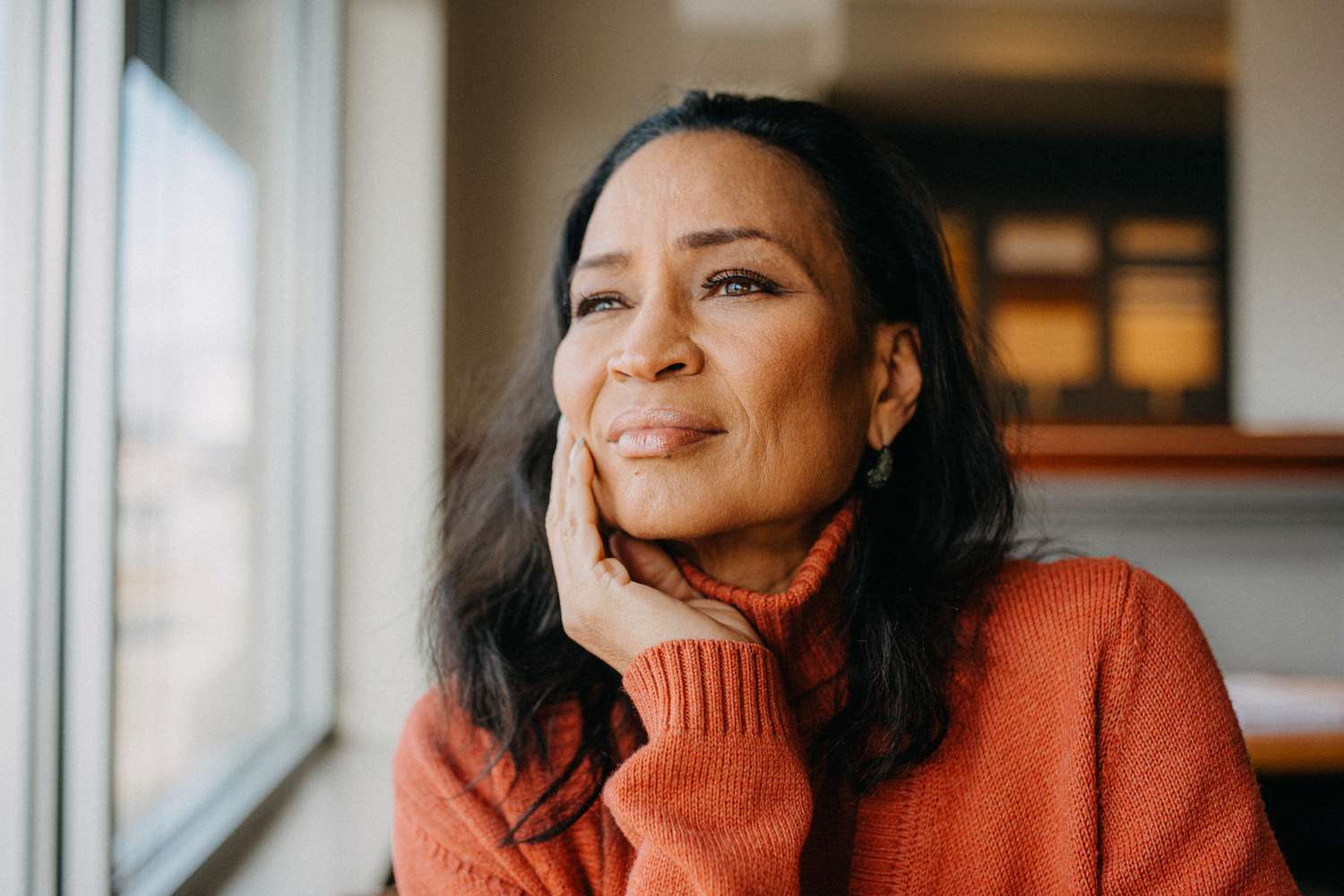 A woman in a cozy orange sweater gazes thoughtfully out of a window, resting her chin on her hand