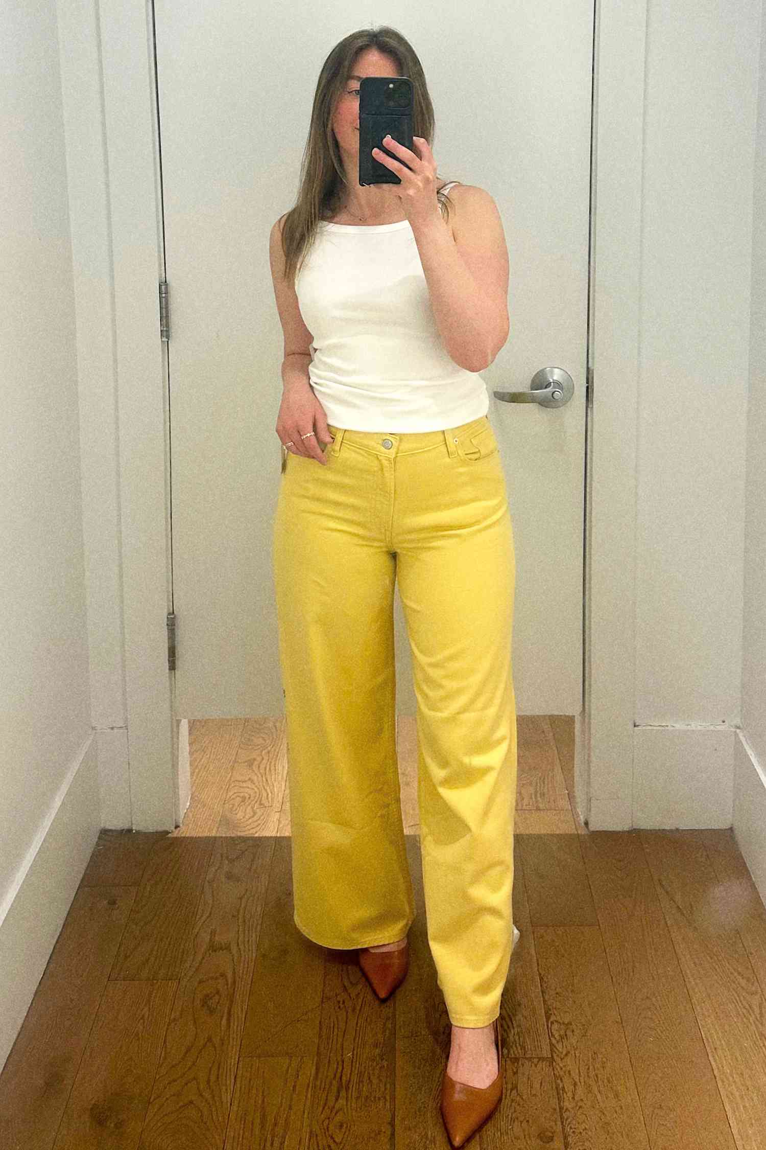 A person taking a selfie in the fitting room wearing a white top and yellow pants