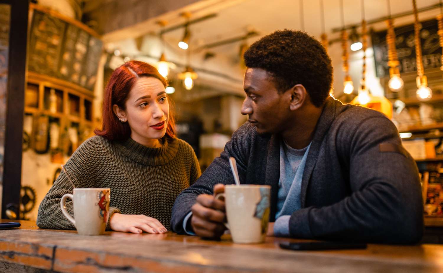 Two individuals sitting at a counter in a café, having a conversation, each holding a mug