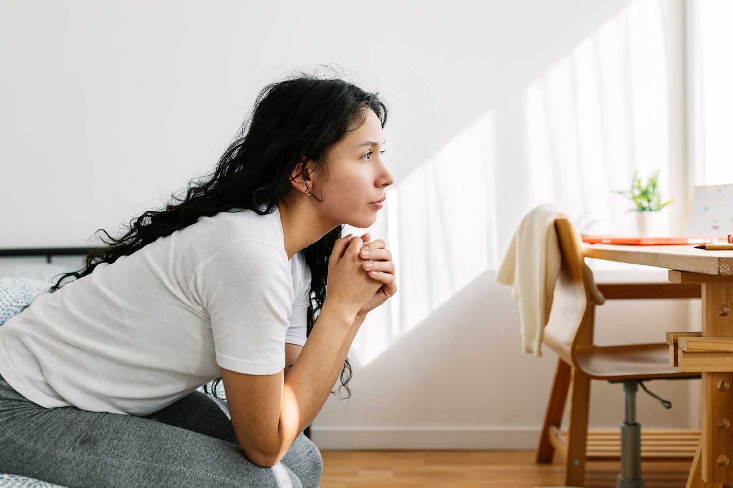 A person sitting on a chair, facing to the right, hands clasped, with a contemplative expression