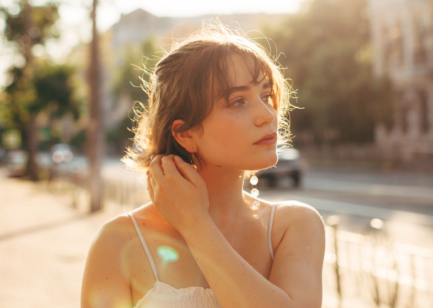 A person standing outdoors, touching their hair, sunlight creating highlights around them in an urban outdoor setting