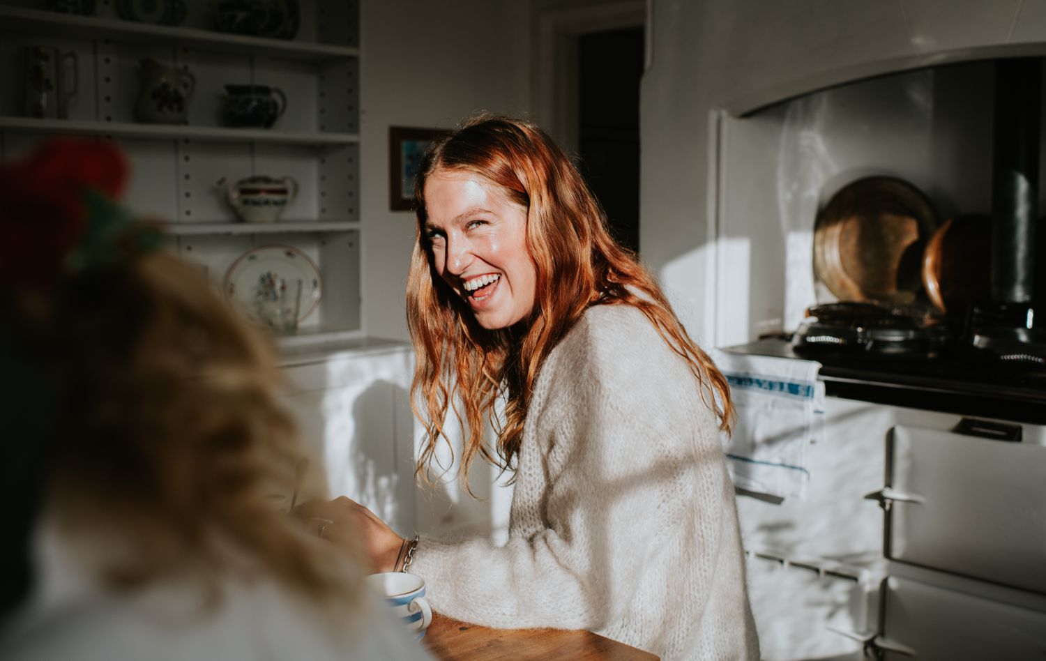 A person indoors smiling while seated at a table in a kitchen setting