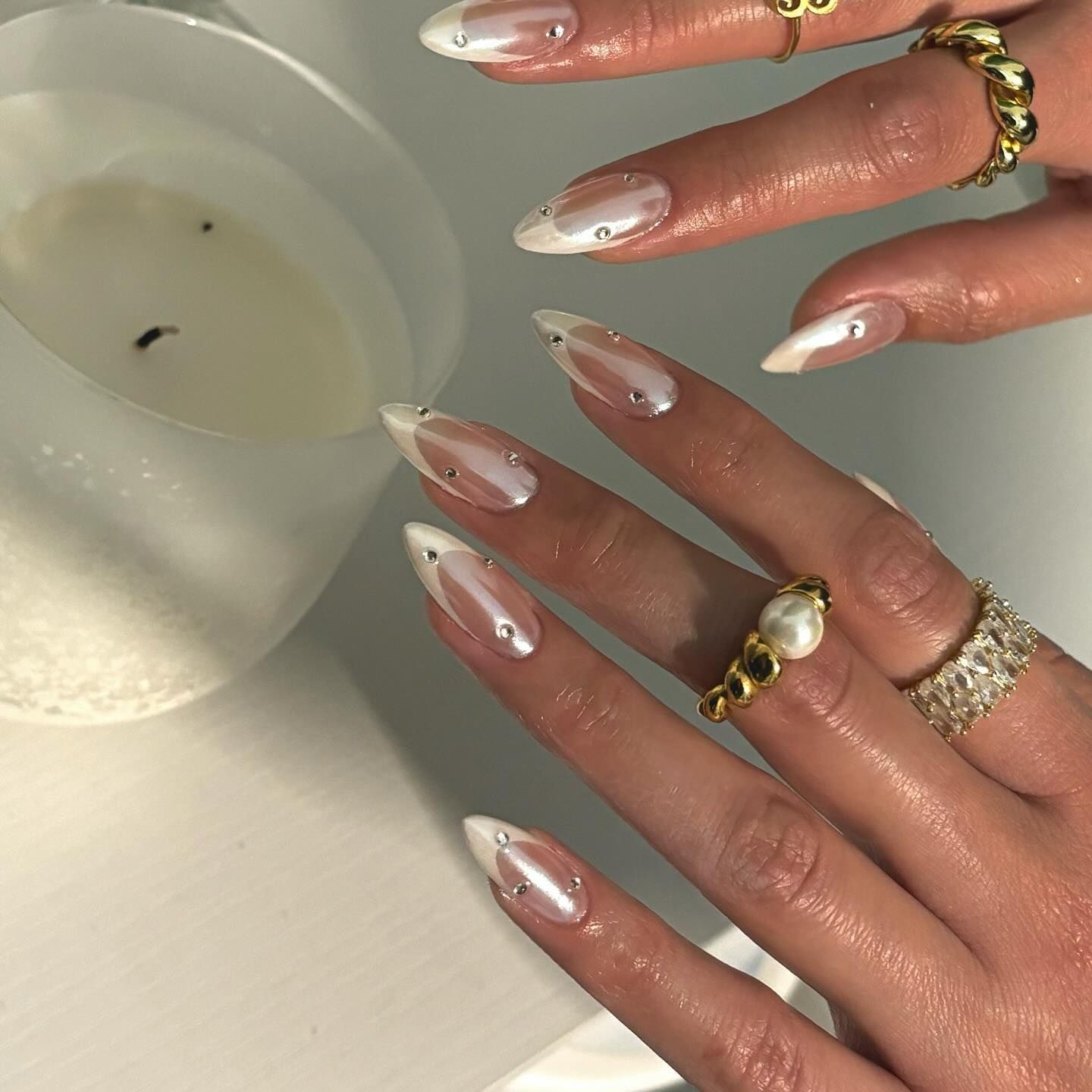 Hands wearing rings with manicured nails near a lit candle, nails have a pearl-like embellishment