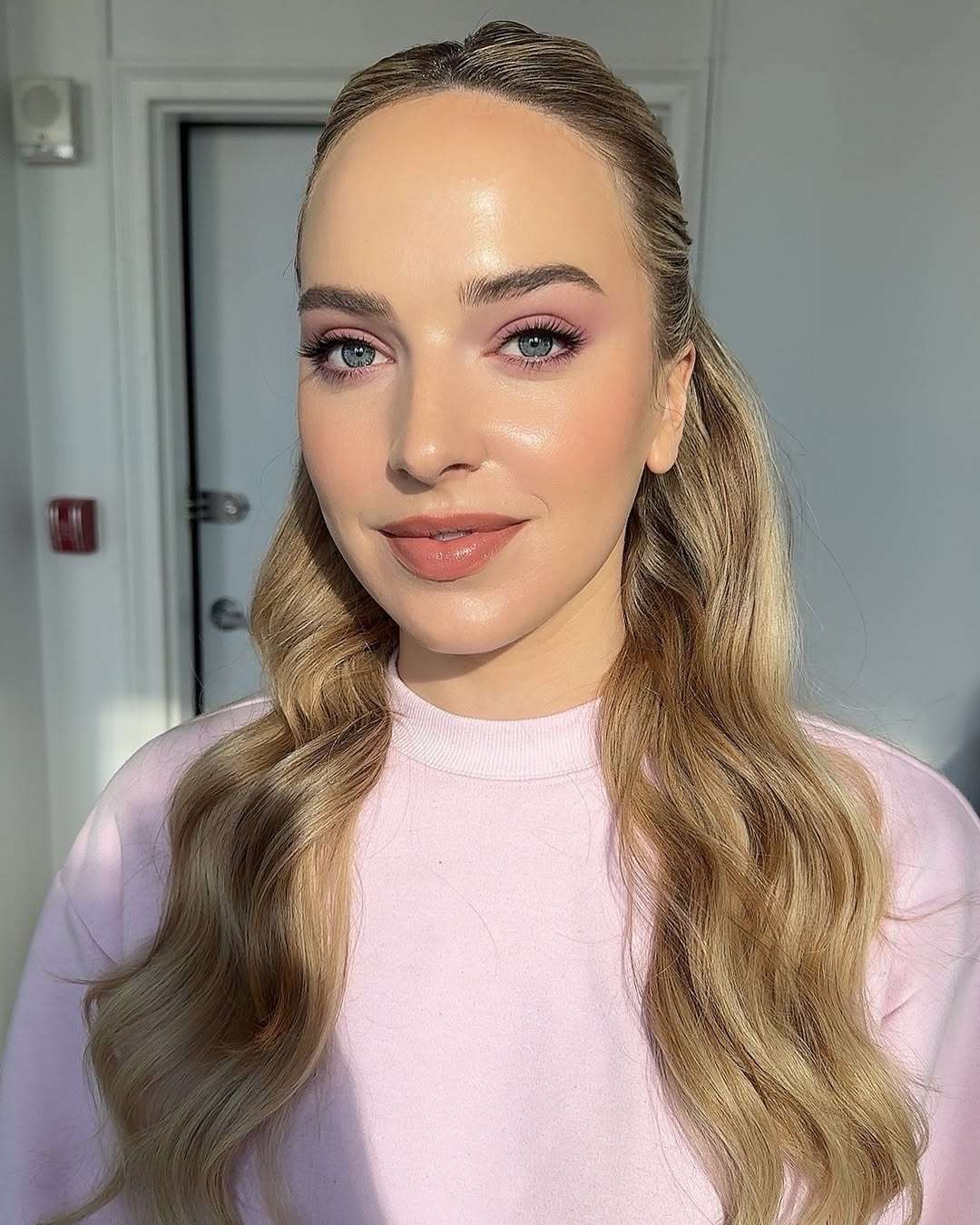 A person with long wavy blonde hair wearing a pink top and pink eyeshadow 