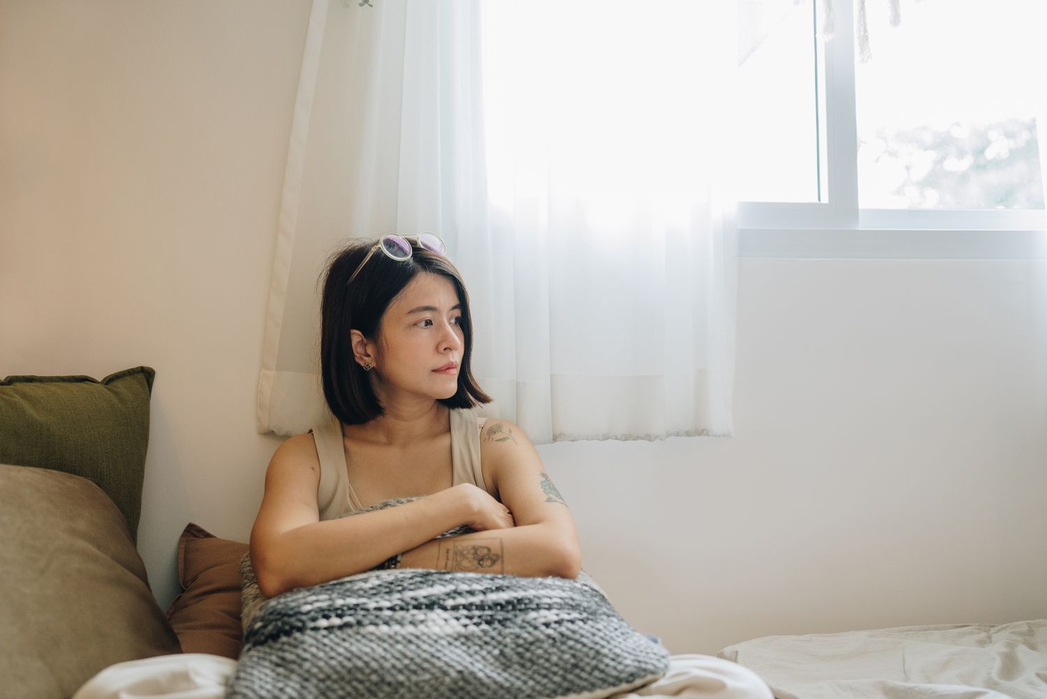 A person sitting indoors next to a window with light streaming through curtains looking thoughtfully to the side
