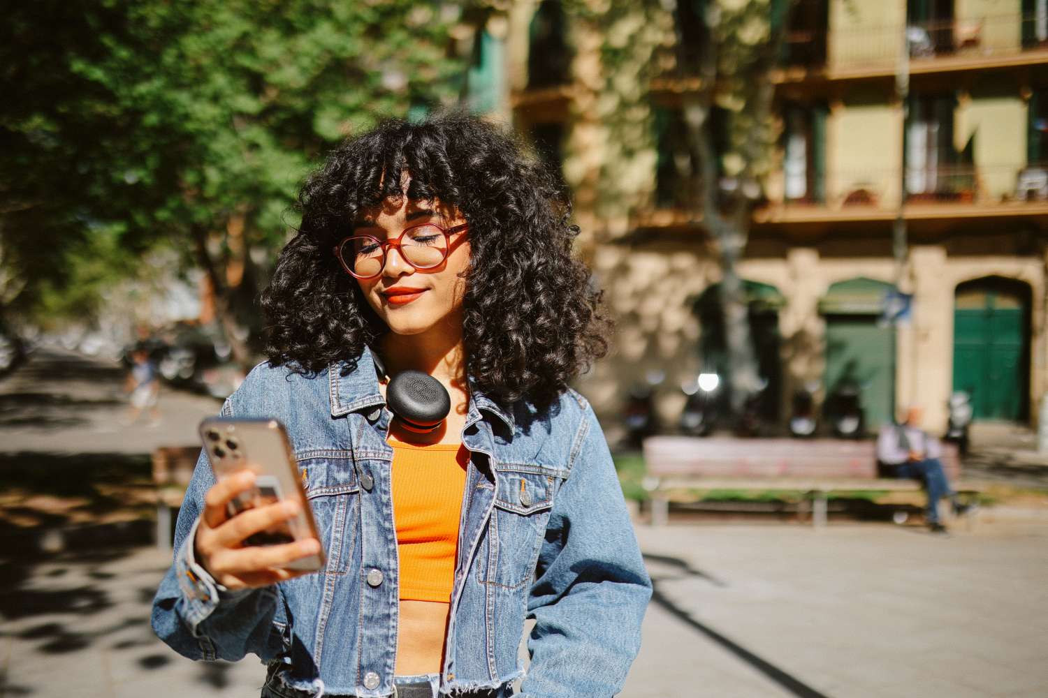 Fashionable young woman with curly hair and glasses stands on a street in Barcelona, wearing headphones and a denim jacket, checking apps on her smart phone.