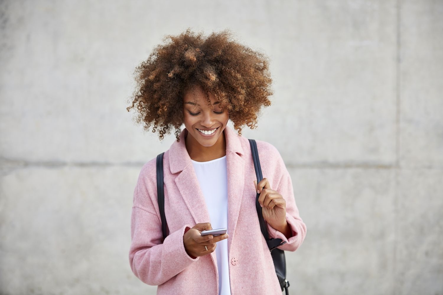 A person wearing a coat smiling and looking at their phone standing in front of a plain wall