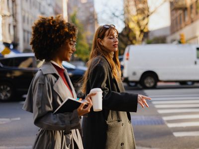 Two individuals walking across a street one holding a notebook and the other a coffee cup with vehicles and buildings in the background