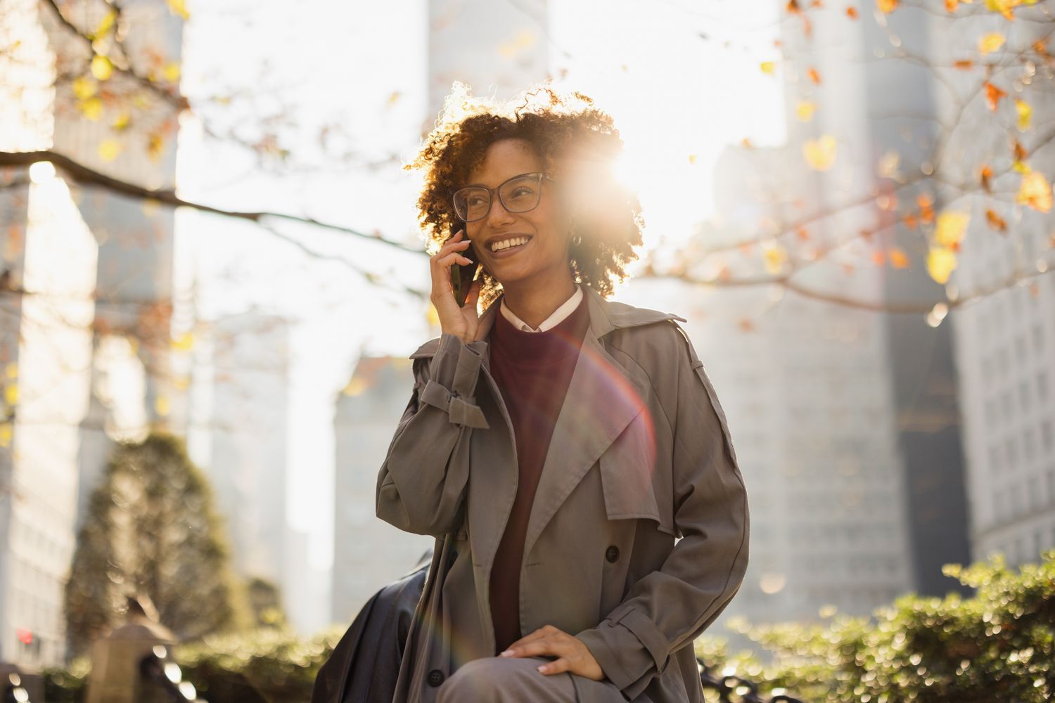 A stylish businesswoman enjoys a sunny day in New York City, smiling while talking on her phone amidst a vibrant cityscape. Her confidence and joy are illuminated by the warm sunlight.