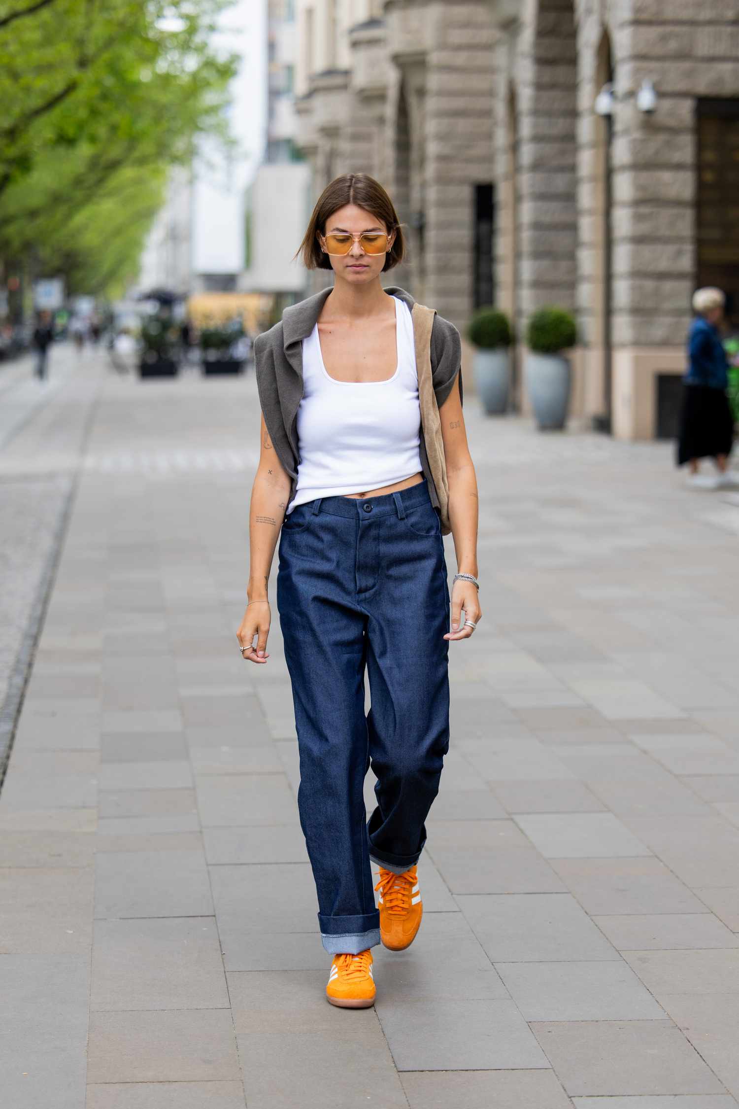 Person with short, brown hair wears orange sunglasses, a white tank, jeans, and orange sneakers