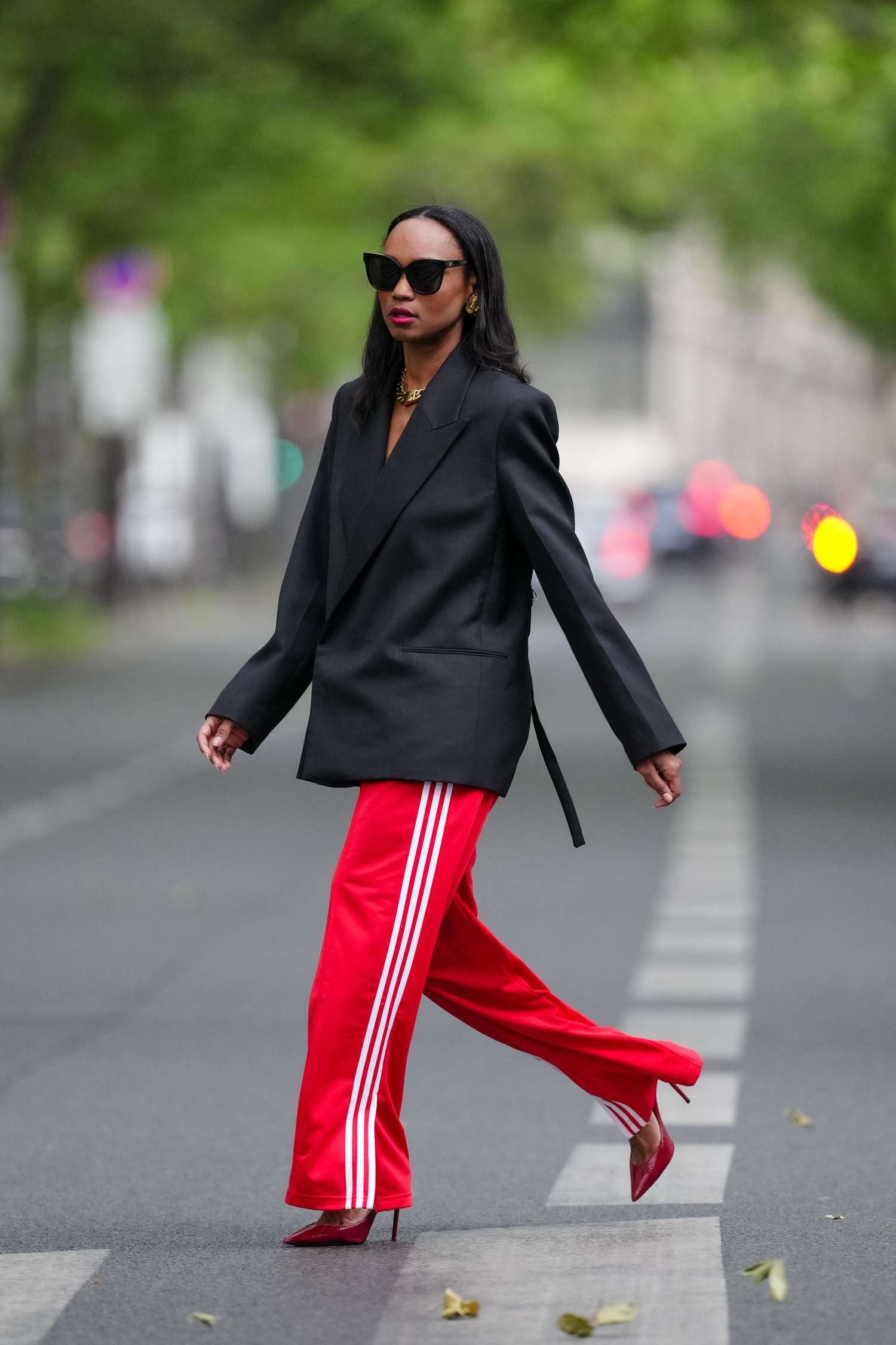 Emilie Joseph wears black sunglasses, oversized black blazer, red Adidas track pants, and red heels