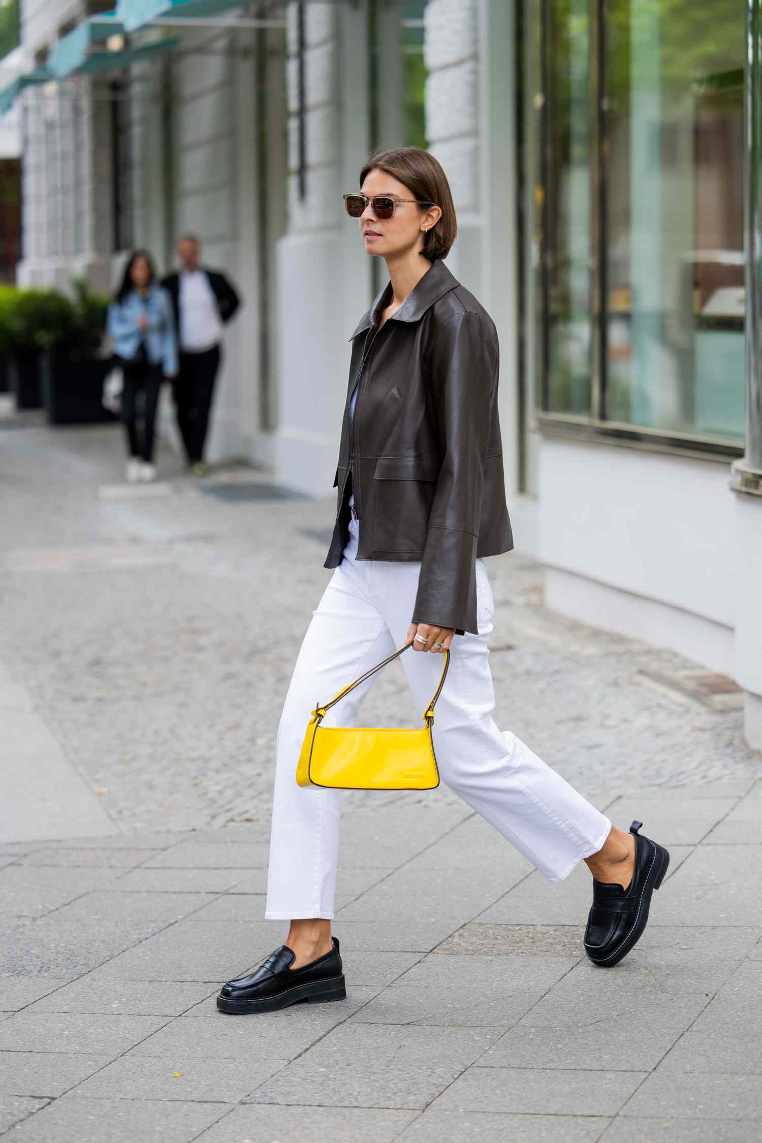 Person with short, brown hair wears a leather jacket, a yellow bag, white pants, and black loafers