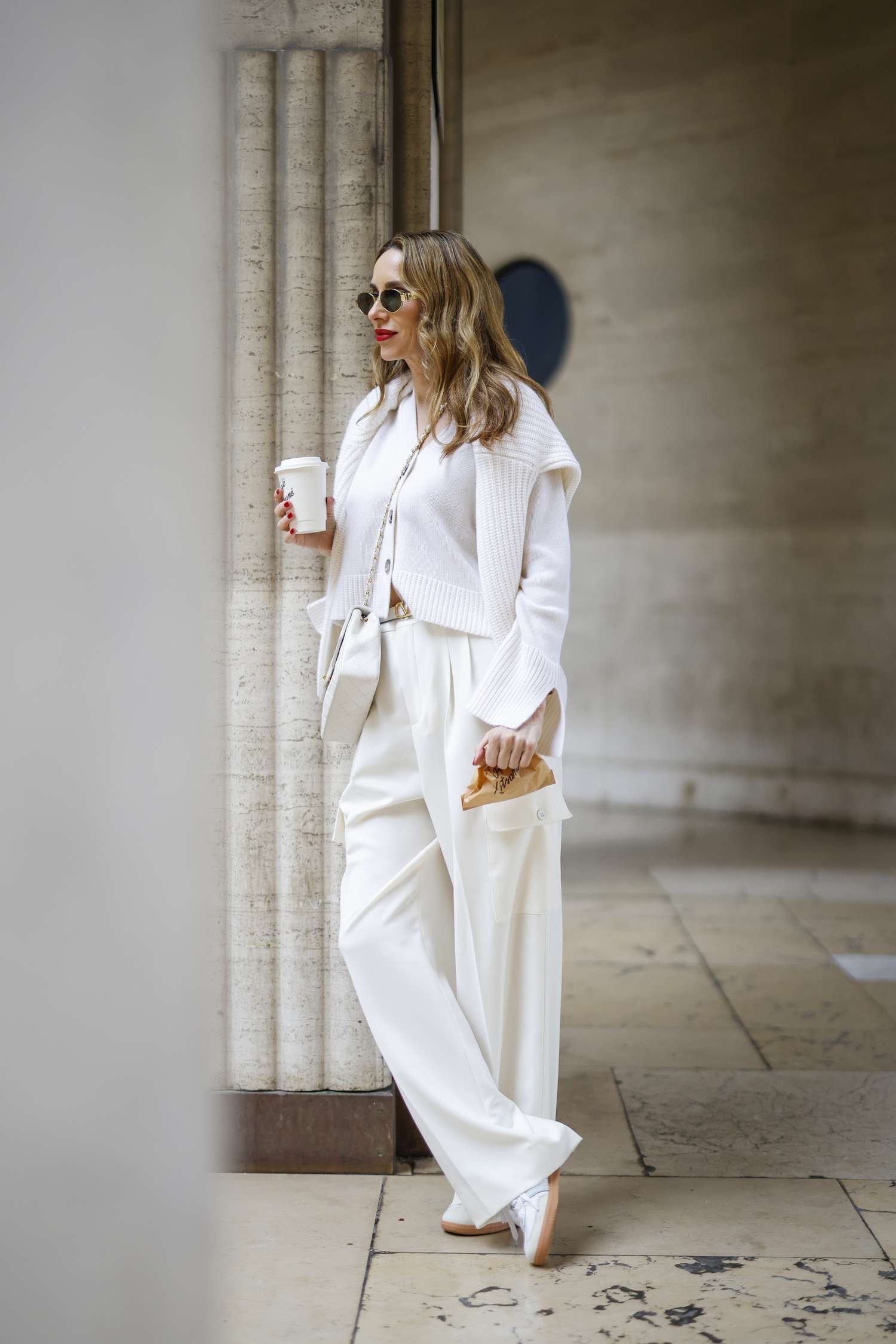 Blonde person with their wavy hair down wears an all-white outfit while holding a white cup