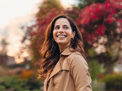Woman with brunette hair wearing gold hoops and a beige trench coat outside during the fall