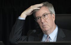 A man with grey hair and glasses with one hand on his head.
