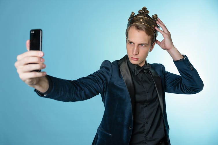 A young man has received an achievement and takes a selfie wearing a suit with a crown on his head.