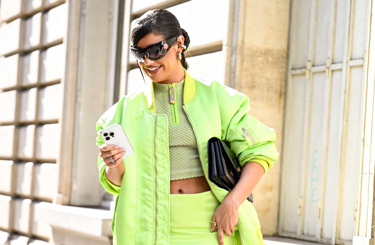 A woman wearing sunglasses and fashionable clothing holding a smartphone and a clutch while outdoors