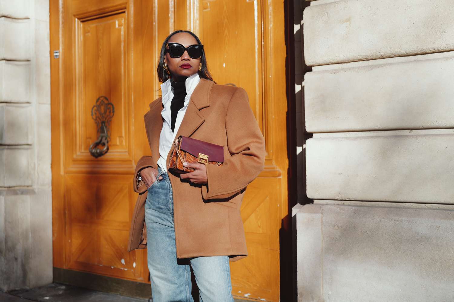 A person standing in front of a wooden door wearing a coat sunglasses and holding a purse while posing