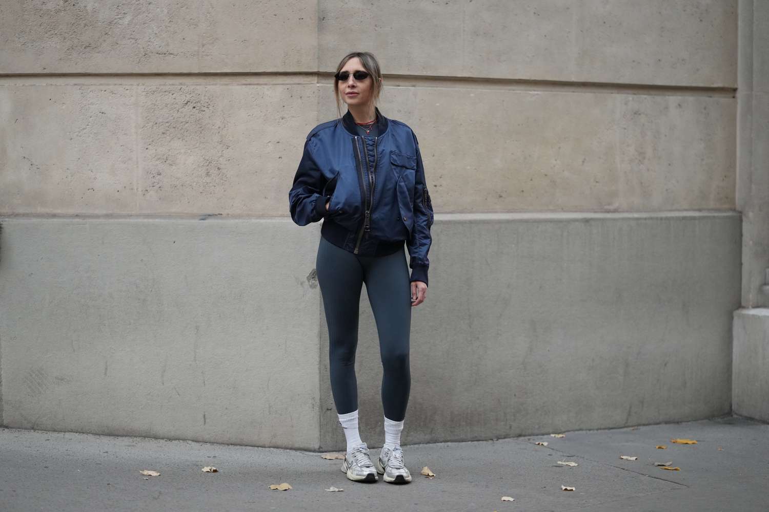 A person wearing a bomber jacket with leggings and sneakers standing against a stone wall sidewalk background