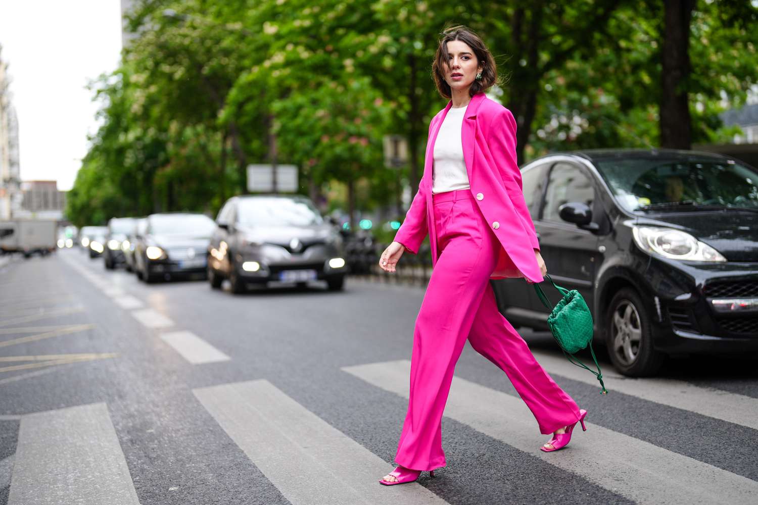 A woman wearing a bright suit walking across a city street while carrying a small handbag