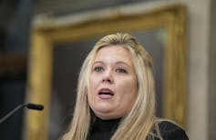 A round-faced blonde woman speaks into a microphone.