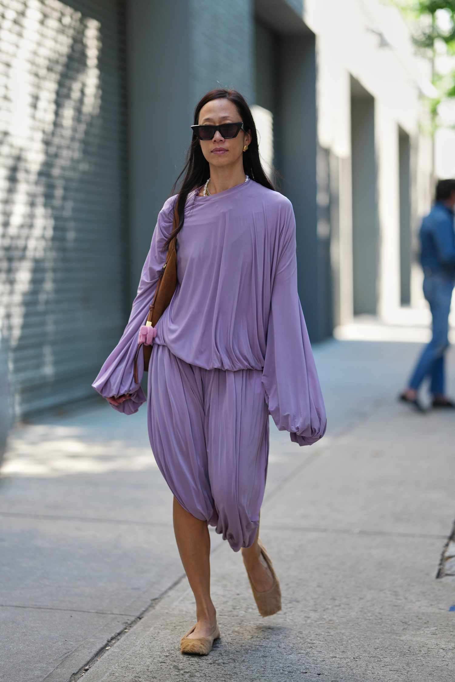 A woman walks along a street wearing a loose outfit and sunglasses carrying a bag